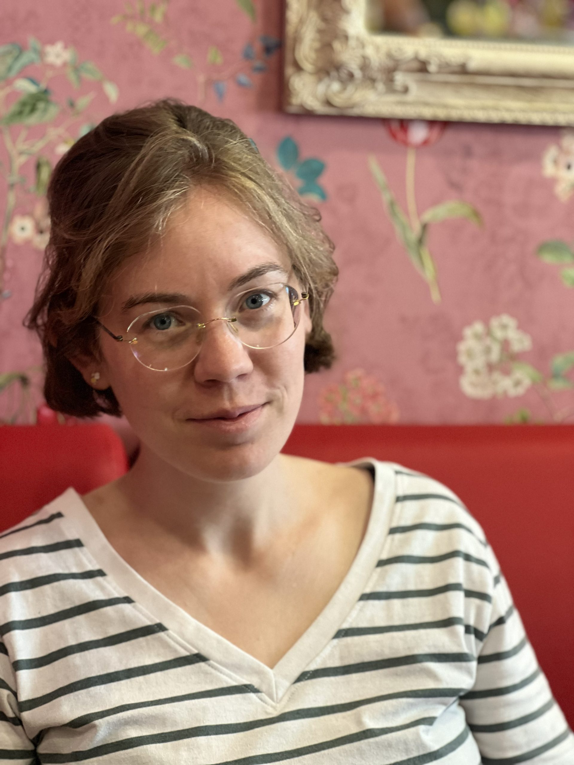 Das bin ich: eine junge Frau mit blonden kurzen Haaren und Brille. Hier trage ich ein Ringelshirt und sitze auf einer roten Bank vor einer rosa Tapete